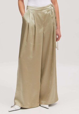 Beige Satinhose mit weitem Bein, vorderen Falten und seitlicher Bindeband-Detail, kombiniert mit weißen spitzen Schuhen und einem weißen ärmellosen Oberteil.