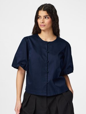 Femme aux cheveux bouclés foncés portant un chemisier bleu marine à manches courtes avec boutons sur le devant et un pantalon noir plissé, debout devant un fond uni.