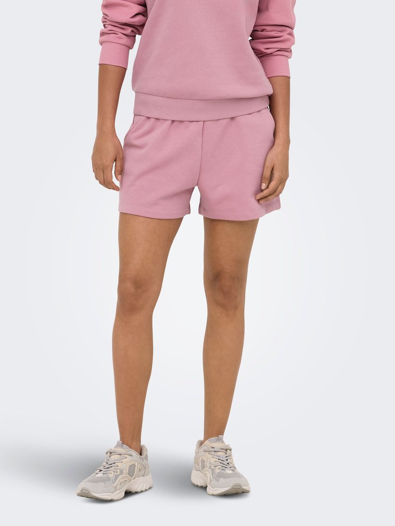 Personne portant un sweat-shirt rose et un short assorti, associés à des chaussures de sport gris clair, se tenant devant un fond clair uni.