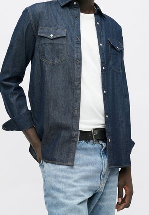 Dunkelblaue Jeanshemd mit Knöpfen, offen getragen über einem weißen T-Shirt, kombiniert mit hellblauen Jeans und schwarzem Gürtel, Hände teilweise in den Taschen.