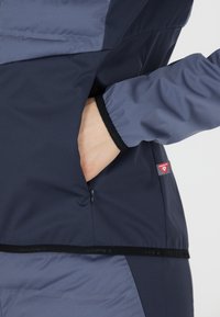 Marineblå og lyseblå atletisk jakke med en lynlås lomme, elastiske cuffs og et rødt logo mærke. Har en glat, holdbar stofstruktur.