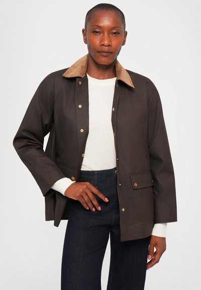 Brown waterproof jacket with a tan corduroy collar, gold snap buttons, and two front pockets, worn over a cream sweater and dark jeans.