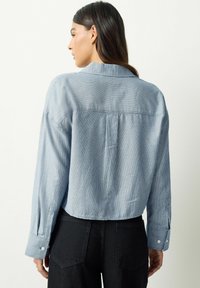 Žena s dlhými tmavými vlasmi, oblečená v modro-bielej pruhovanej košeli na gombíky a čiernych nohaviciach, zobrazená zozadu na jednoduchom pozadí.