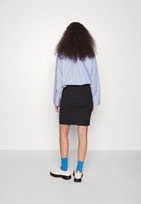 Chemise légère bleu clair, jupe crayon noire, chaussettes bleues et chaussures blanches avec semelles noires, vues de dos sur un fond uni.