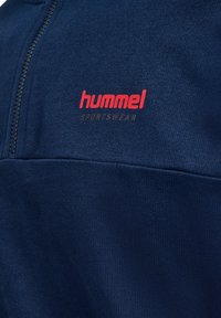 Maglietta sportiva blu navy con colletto alto, dotata di chiusura con zip e logo "hummel" rosso sul petto, realizzata in tessuto testurizzato.