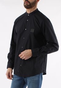 Camicia nera con colletto rialzato, dotata di una piccola toppa con logo sul lato sinistro del petto e bottoni blu a contrasto. Realizzata in tessuto morbido.