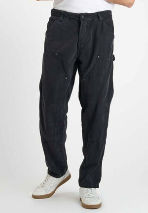Pantalons cargo noirs avec plusieurs poches, coupe décontractée, tissu texturé et fermeture à bouton sur le devant, associés à des baskets blanches.