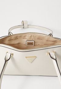 Borsa bianca in materiale sintetico con hardware dorato, dotata di chiusura con zip e interno beige foderato con un motivo ripetuto di logo "GUESS".