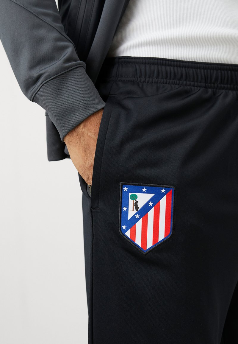 Un pantalon de sport noir présente un emblème aux couleurs bleu, rouge et blanc, représentant un écu avec des étoiles et un design en silhouette.