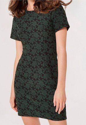 Vrouw in een zwart jurkje met korte mouwen en tot de knie, met een donkergroen bloemenpatroon, staand tegen een lichte achtergrond.