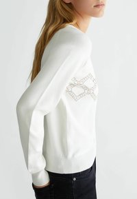 Maglione bianco a maglia con polsini e orlo a coste. Presenta un motivo geometrico accentuato da strass sul petto per un tocco di dettaglio in più.