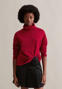 Roter Rollkragenpullover mit strukturierter Oberfläche, kombiniert mit einem schwarzen Cordrock mit Falten. Einfaches Design, mittellange Ärmel.