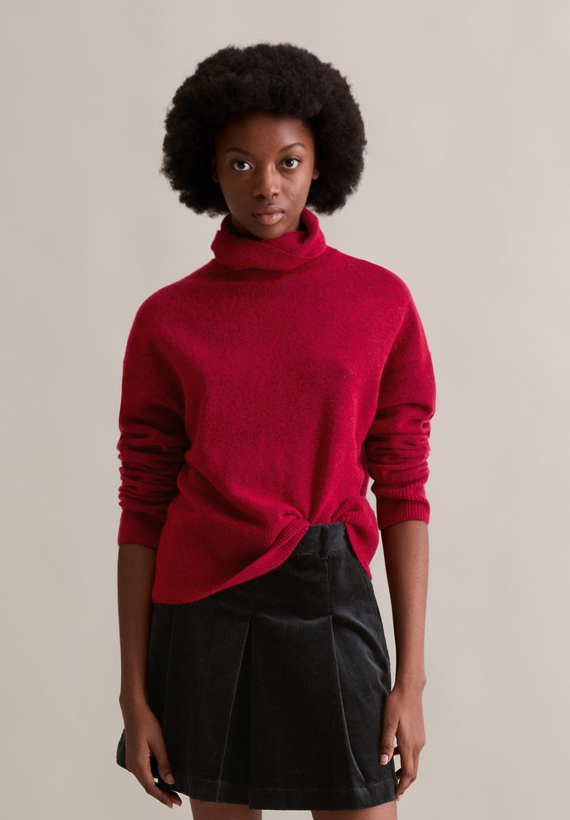 Roter Rollkragenpullover mit strukturierter Oberfläche, kombiniert mit einem schwarzen Cordrock mit Falten. Einfaches Design, mittellange Ärmel.