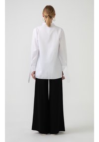 Witte blouse met lange mouwen, hoge kraag en brede manchetten, gedragen met zwarte wijde broek, tegen een effen achtergrond.