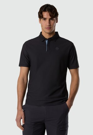 Uomo giovane con capelli corti e scuri, che indossa una polo nera a maniche corte e pantaloni scuri, in piedi davanti a uno sfondo grigio chiaro uniforme.