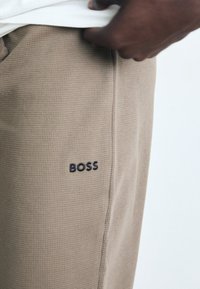 Beige Strick-Jogginghosen mit einer strukturierten Oberfläche und einem kleinen marineblauen "BOSS"-Logo auf der linken Seite. Verfügt über einen entspannten Schnitt und einen elastischen Bund.