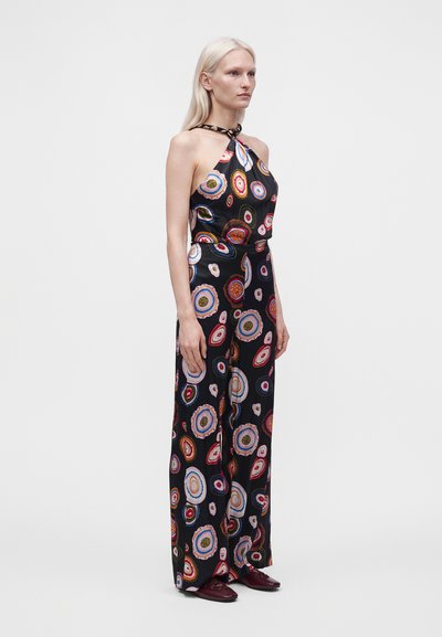 Femme aux longs cheveux blonds portant une combinaison noire halter avec des motifs circulaires colorés et des chaussures bordeaux, debout devant un fond uni.