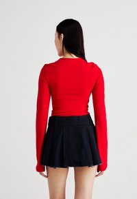 Haut rouge à manches longues en tissu stretch ajusté, associé à une mini-jupe plissée noire avec un ourlet effiloché.