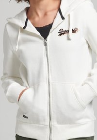 Sudadera blanca con cremallera hecha de un tejido suave, que cuenta con una capucha con ribete negro, bordado del logo y bolsillos frontales prácticos.