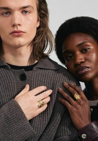 Dos personas llevan suéteres oscuros y texturizados, mostrando grandes anillos metálicos con diseños intrincados y acabados contrastantes en sus manos.