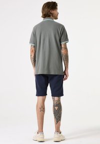Grijze polo met lichtblauwe accenten, gecombineerd met marineblauwe shorts. Heeft korte mouwen, een relaxte pasvorm en zichtbare tatoeages op de benen.