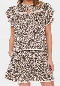 Kobieta ubrana w bluzkę z bufkami w panterkę i pasujące szorty z koronkowym wykończeniem, z rękami wsuniętymi w kieszenie, na białym tle.