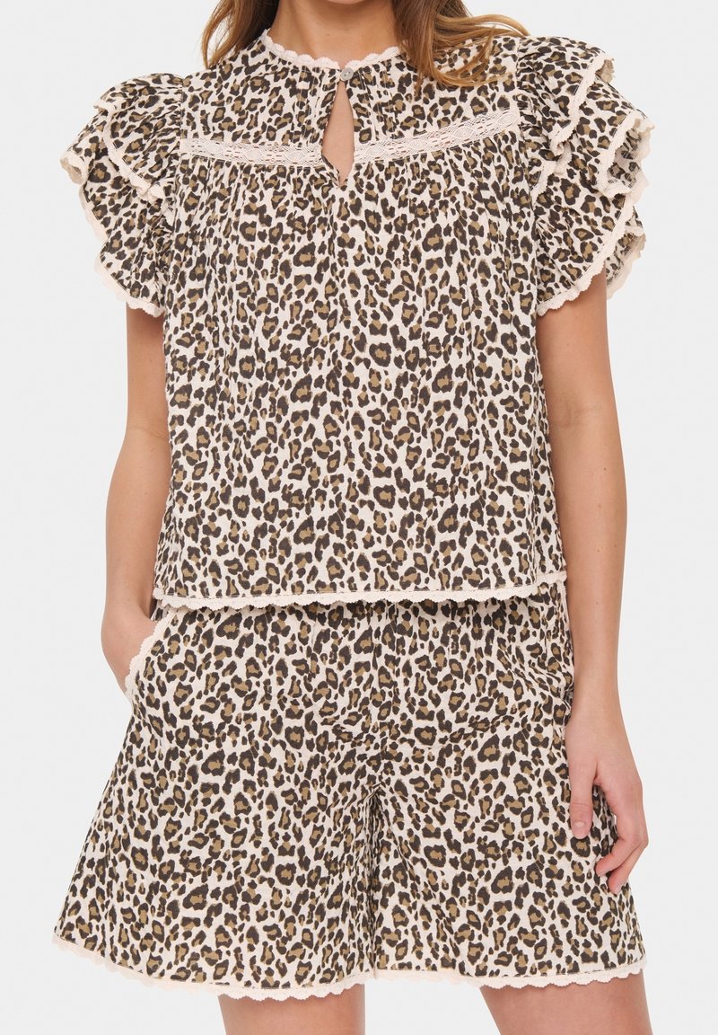 Kobieta ubrana w bluzkę z bufkami w panterkę i pasujące szorty z koronkowym wykończeniem, z rękami wsuniętymi w kieszenie, na białym tle.