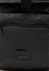 Crni najlon ruksak s prednjim zip džepom, podešavanim trakama i teksturiranim logom. Glatka površina s elegantnim dizajnom.