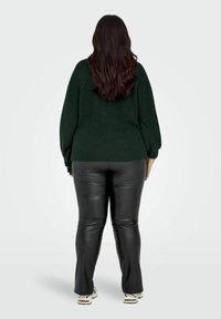 Tmavě zelený pletený svetr s volnými rukávy, kombinovaný s černými lesklými kalhotami z umělé kůže a světlými sportovními botami. Pohled zezadu.