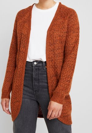 Mujer con un cárdigan de punto grueso color óxido abierto al frente, sobre una camiseta blanca y unos vaqueros negros de cintura alta.