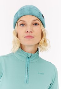 Šviesiai mėlynas riebus kepurė su etikete, derinama su šviesiai mėlynu užtrauktuko kaklo viršumi, kuriame yra atspaudas. Minkšta tekstūra ir prigludęs dizainas.