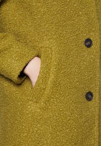 Olivgrön texturerad kappa i mjukt, fluffigt material, med en stor sidoficka och svarta knappar framtill.