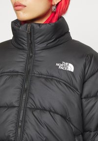 Svart vadderad jacka med hög krage, horisontell quiltad mönster, dragkedja framtill och en vit logotyp på vänster bröst.