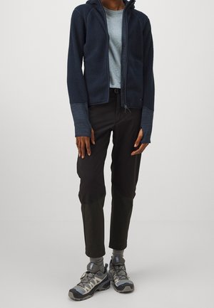 Veste polaire marine avec capuche, chemise bleu clair, pantalon de randonnée noir et chaussures de randonnée grises avec accents foncés et semelles texturées.