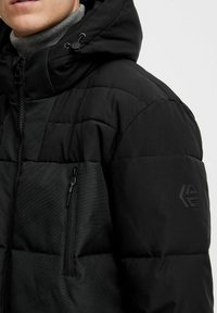 Sort puffjakke med en struktureret finish, der har en lynlåslomme og hætte med snøre. Akzentueret med et diskret logo på ærmet.