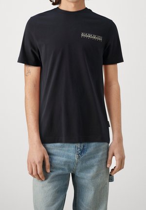 Mand iført en sort t-shirt med korte ærmer med et lille "Napapijri geographic" logo på brystet og lyseblå jeans.