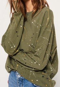 Sudadera verde oliva con patrones de salpicaduras de pintura abstracta en blanco, amarillo y negro. Presenta un corte relajado y detalles desgastados.