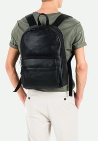 Sac à dos en cuir noir avec une texture lisse, un haut arrondi, une poche zippée à l'avant, des bretelles ajustables et une boucle de suspension sur le dessus.