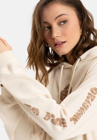 Sudadera beige con cordón, con un logo blanco y texto en marrón en la manga. Tela suave con un corte holgado.