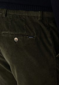 Pantaloni in velluto a coste verde con una tasca posteriore dotata di un bottone argento e una piccola etichetta. Il tessuto presenta un aspetto testurizzato e rigato.
