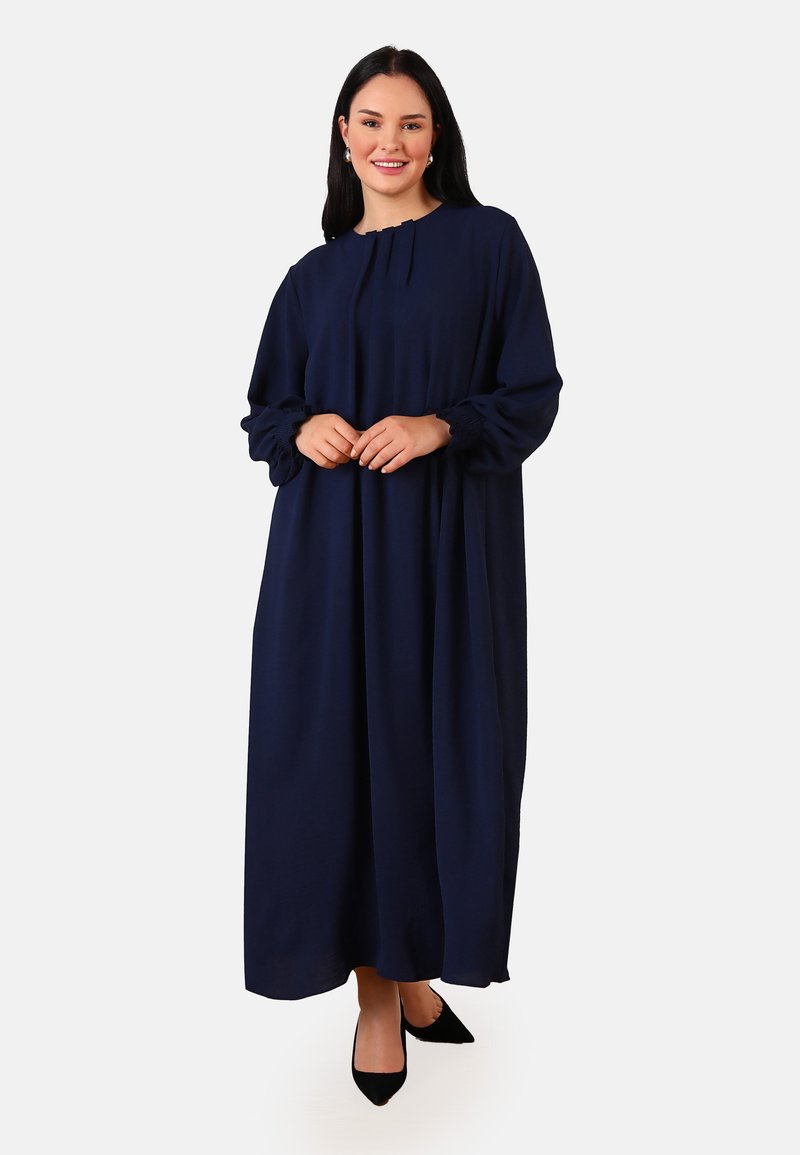 Robe longue à manches longues, bleu marine, en tissu doux avec un design plissé et des poignets élastiques. Accompagnée de chaussures noires à bout pointu.