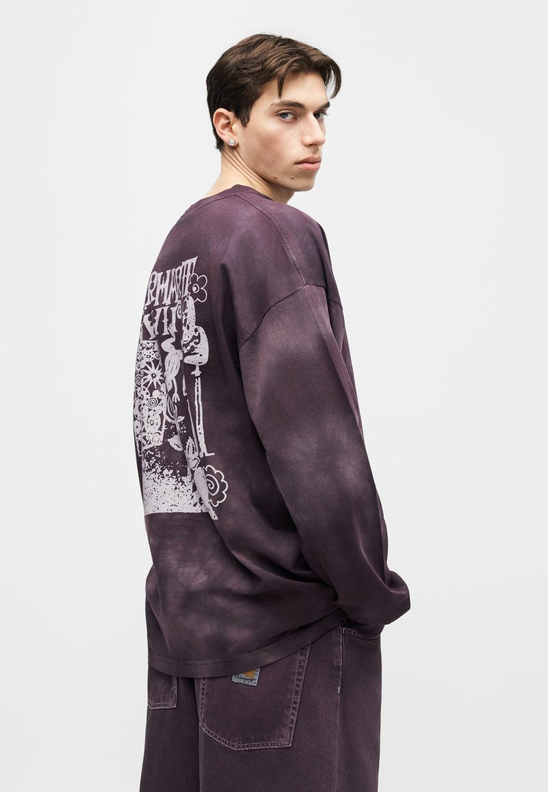 Giovane uomo che guarda oltre la spalla indossando una maglietta a maniche lunghe tinta in capo viola con grafica astratta bianca e pantaloni abbinati su sfondo bianco.