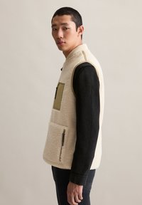 Jonge man in een crèmekleurige fleece vest met olijfgroene zakken over een zwart T-shirt met lange mouwen, staande in profiel tegen een effen lichte achtergrond.