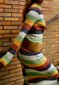 Femme portant une robe rayée multicolore avec une ceinture marron montant des escaliers devant un mur en briques.