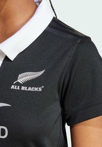 Svart rugbytröja med vit krage, har "ALL BLACKS"-logotypen och silverfjäderemblemet, tillverkad av textilerat, andningsbart material.
