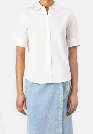 Henkilö pukeutuneena valkoiseen lyhythihaiseen napitettuun paitaan ja vaaleansiniseen farkkuun kietaisuhameeseen, seisomassa yksiväristä taustaa vasten.