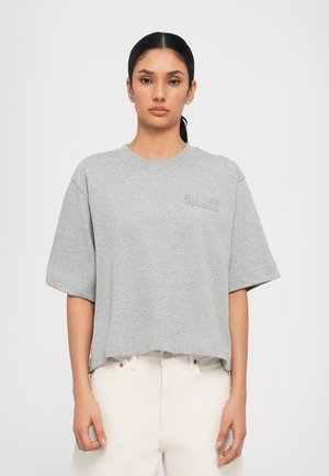 Grijze katoenen t-shirt met korte mouwen, ronde hals en subtiele branding op de borst. Gedragen met lichtgekleurde broek.