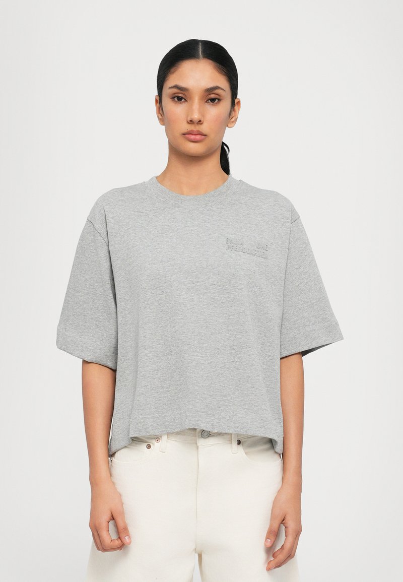 Camiseta de algodón gris de manga corta, cuello redondo y con una sutil marca en el pecho. Llevada con pantalones de color claro.