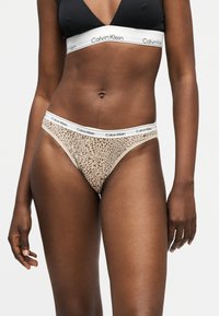 Γυναίκα που φοράει μαύρο μπραλέτο Calvin Klein και εσώρουχο με λεοπάρ σχέδιο Calvin Klein με λευκές ελαστικές ταινίες με το λογότυπο, μπροστά σε απλό φόντο.