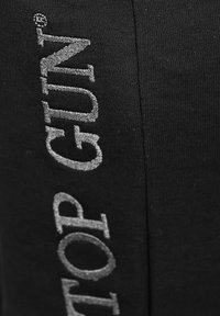 Svart tyg med stora silverbroderade "TOP GUN"-texter, med en slät yta och tät sömnadsdetalj.